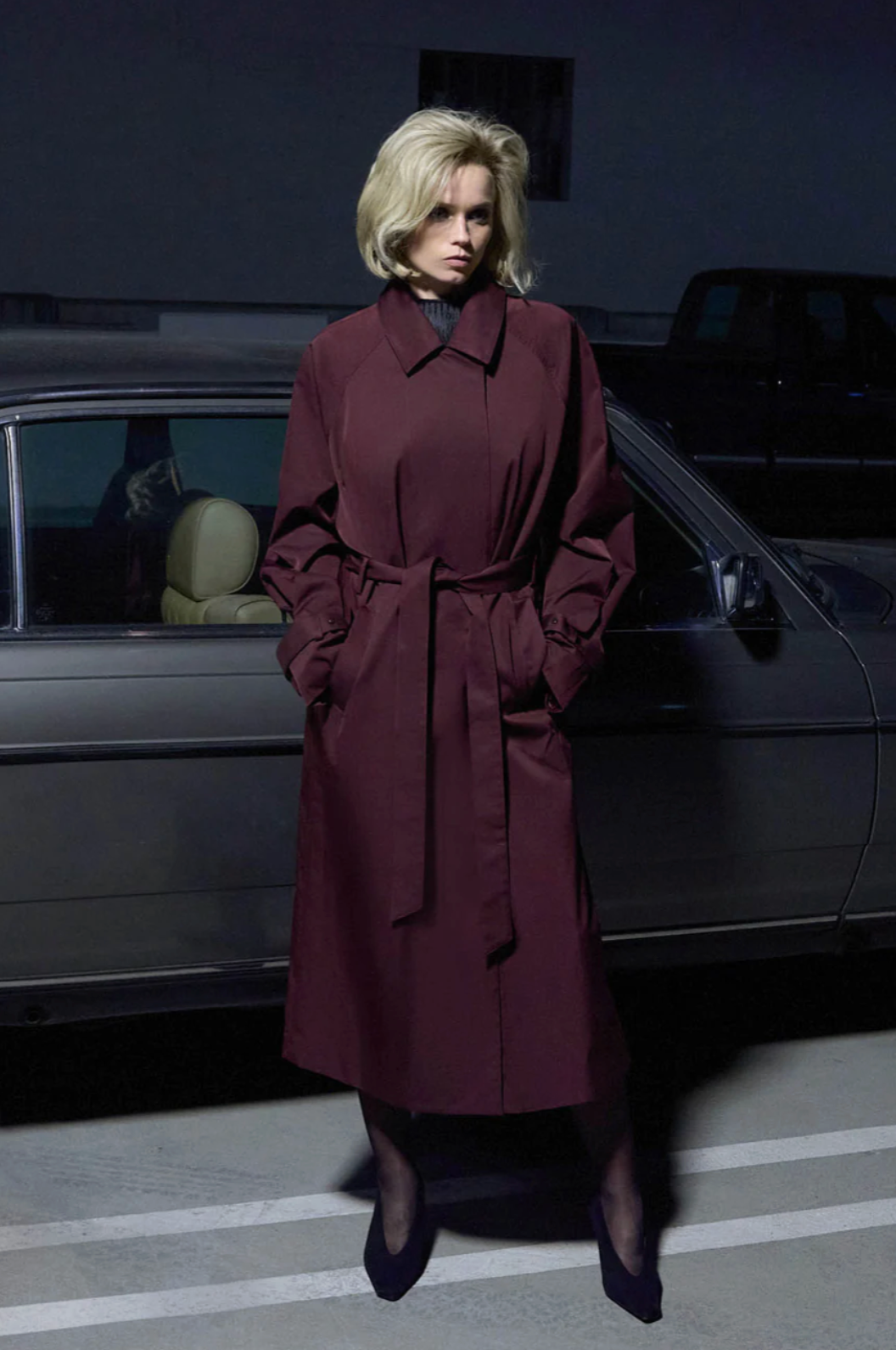 Woman in a long coat standing next to a car in a dimly lit setting