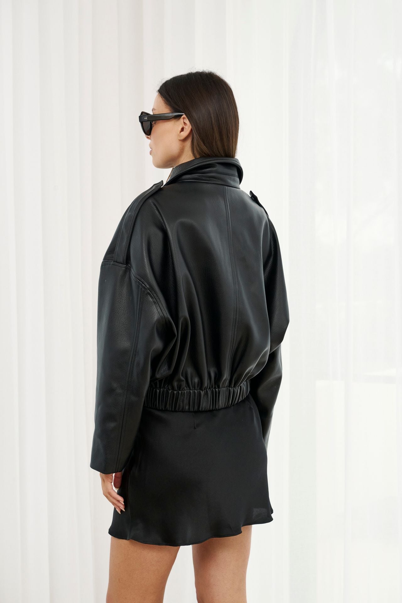 Person wearing a black leather jacket and skirt set against a white curtain background