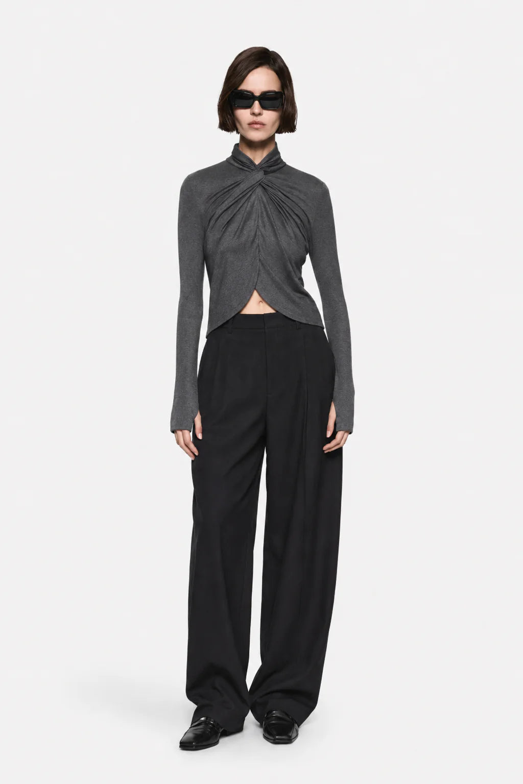 A model wearing a dark gray long-sleeve top with a twisted front detail and black wide-leg trousers, standing against a light gray background.