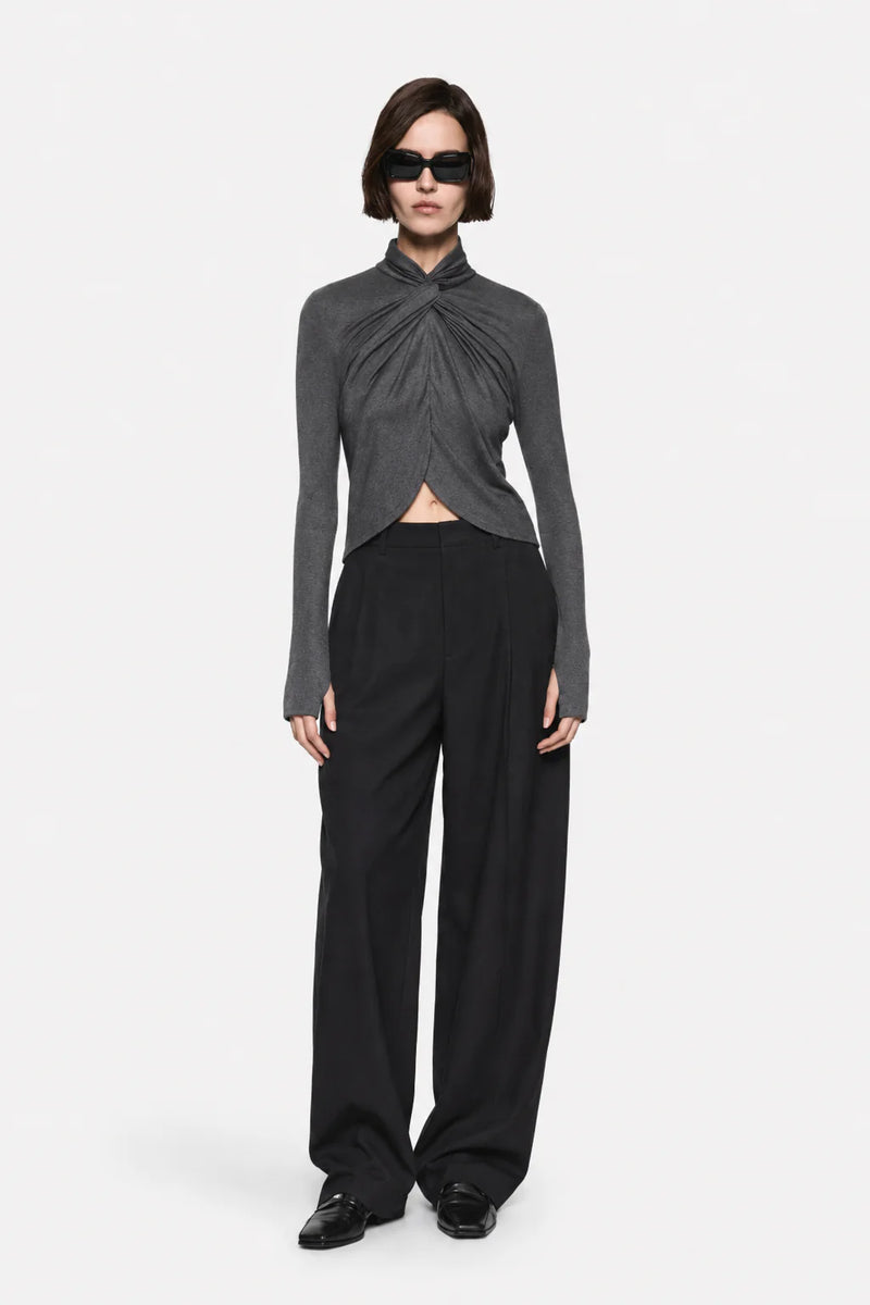 A model wearing a dark gray long-sleeve top with a twisted front detail and black wide-leg trousers, standing against a light gray background.