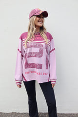 A woman wearing a light pink and dark pink long-sleeve athletic-style sweatshirt with a large distressed number 21 on the front, paired with black pants and a matching pink cap, standing against a plain white wall.