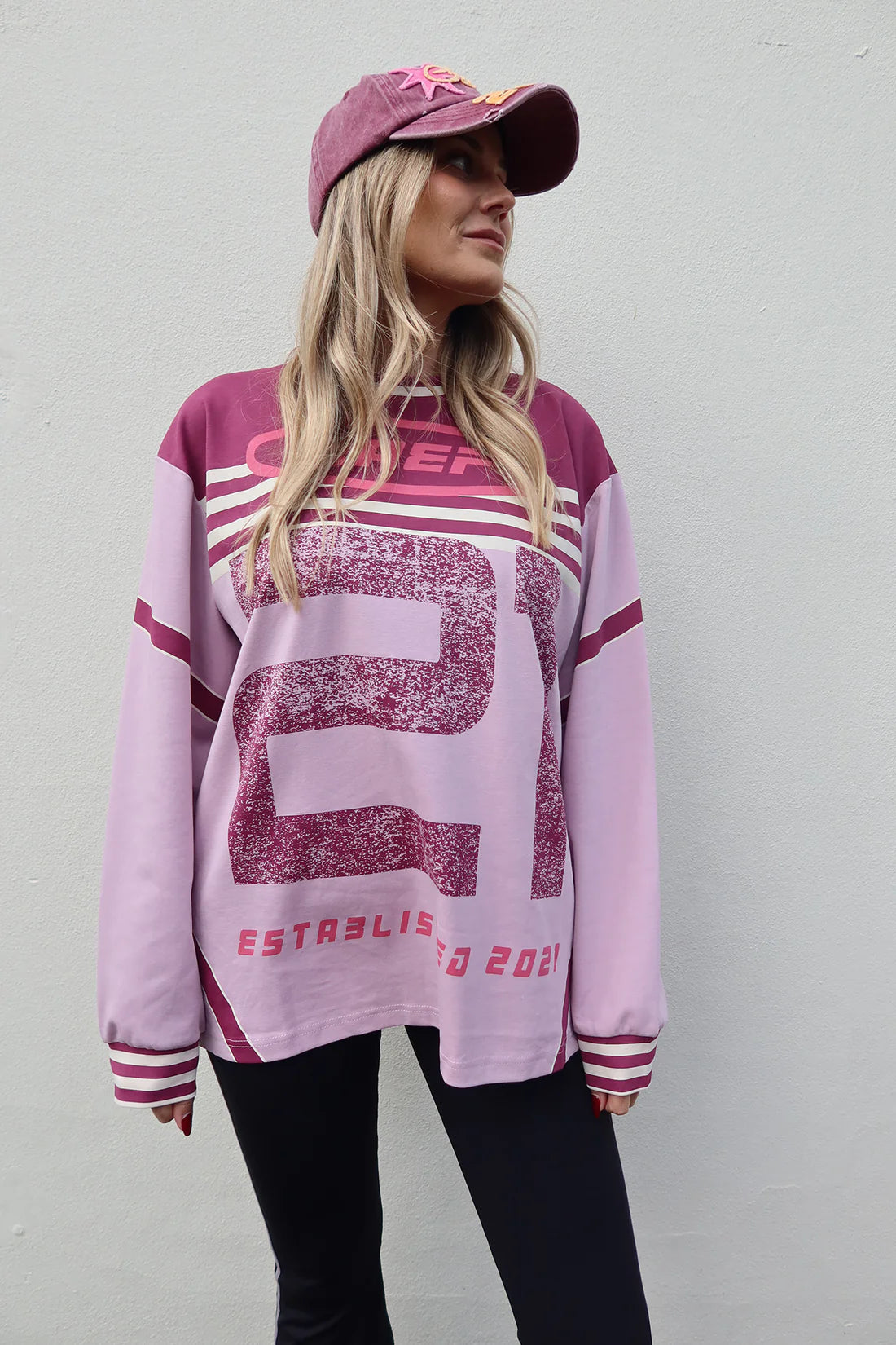 A woman wearing a pink and purple distressed sports jersey sweatshirt, a matching pink baseball cap, and black pants, standing against a light gray wall.