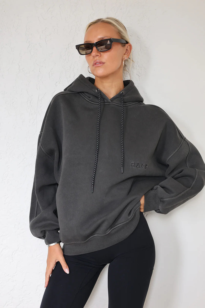 A woman wearing a dark charcoal gray oversized hoodie with black drawstrings and a tonal embroidered logo on the chest, paired with black leggings, posing against a white textured wall.