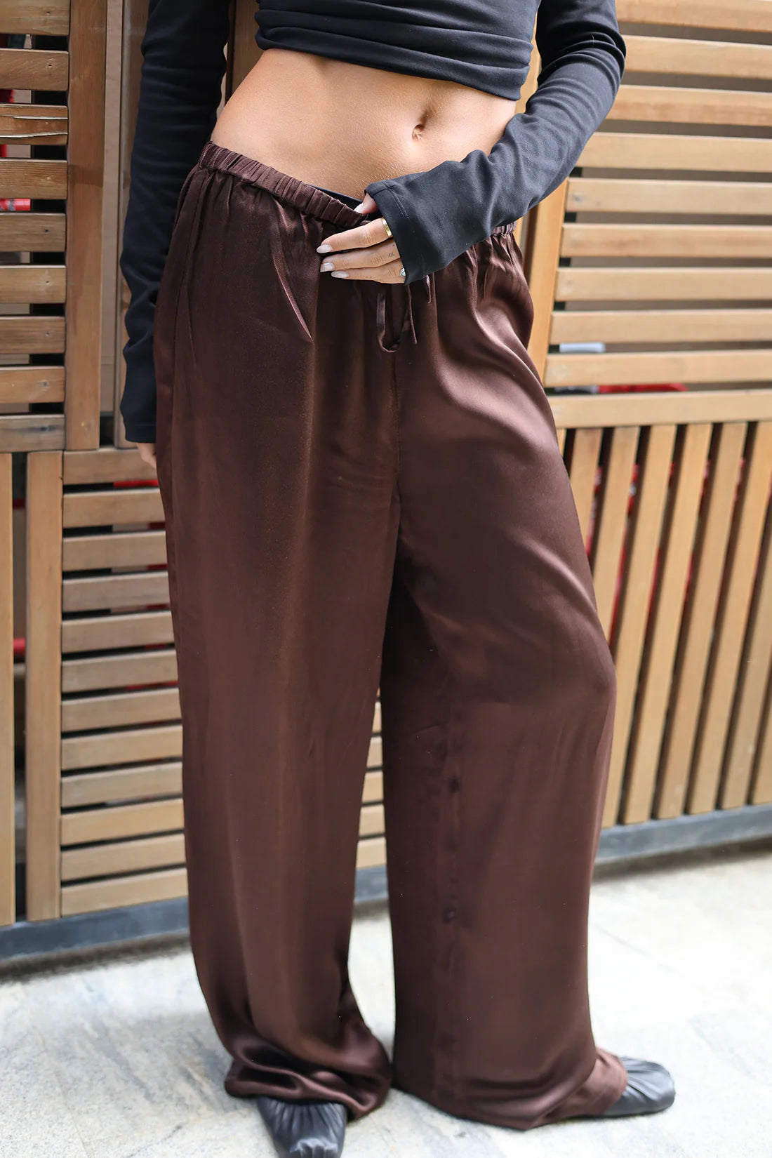 A person wearing loose, wide-leg brown satin or silky pants with an elastic drawstring waist, paired with a dark long-sleeve crop top and black shoes, standing in front of a wooden slat wall.