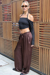 A woman wearing a black off-the-shoulder long-sleeve crop top and brown wide-leg satin pants, standing in front of a wooden slat wall.