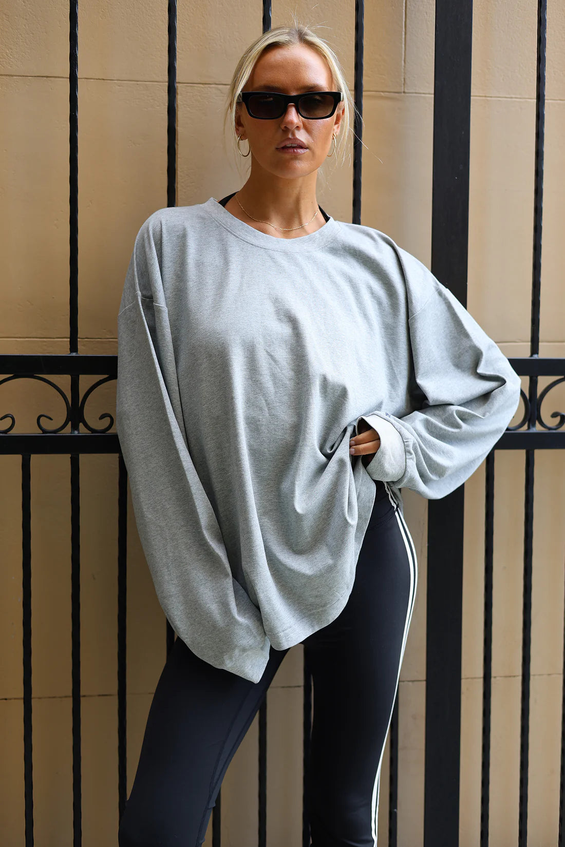 A woman wearing an oversized light gray crewneck sweatshirt and dark leggings with white side stripes, standing in front of a beige wall with black metal bars.