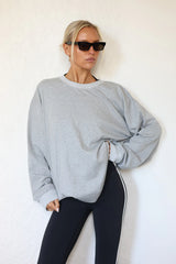A woman wearing an oversized light gray crewneck sweatshirt and black leggings with a white side stripe, posing against a white textured wall.
