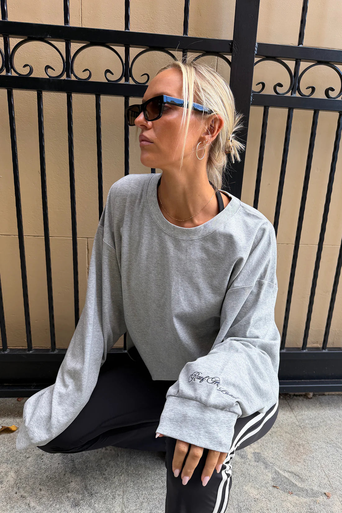 A woman wearing a light gray oversized crewneck sweatshirt, black track pants with white stripes, and sunglasses, sitting in front of a beige wall and black metal fence.