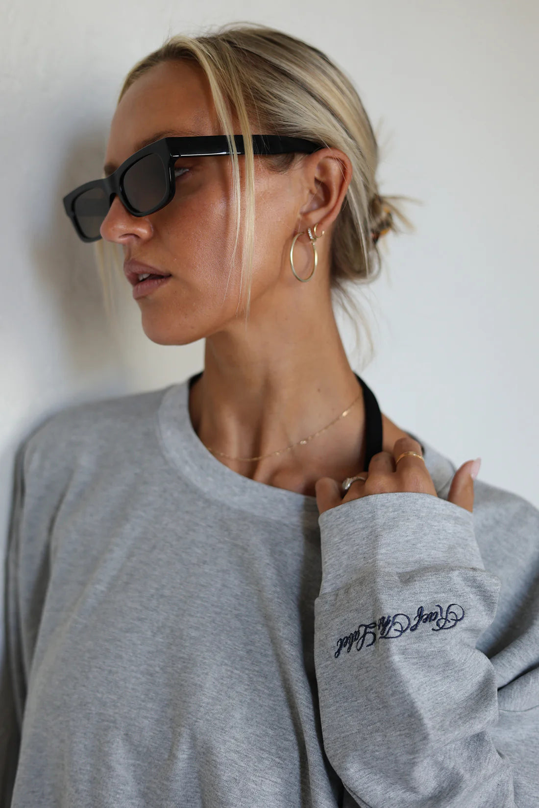 A woman wearing a light gray crewneck sweatshirt with a black embroidered script logo on the left sleeve, styled with black sunglasses and gold hoop earrings against a plain white wall.